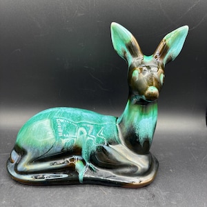 May include: A ceramic figurine of a deer in a resting position, featuring a glossy glaze with blended shades of deep black and vibrant turquoise green. The deer is depicted with its legs tucked underneath its body and ears upright.