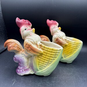 May include: Two ceramic rooster figurines, each holding a cornucopia basket. The roosters have pink combs, white faces, and colorful plumage. The baskets are yellow and green, with a purple base. The background is black.