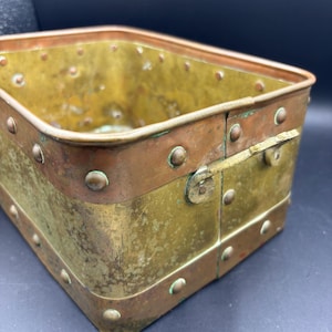 May include: A rectangular metal container with a brass body and copper trim. The container is riveted together with small, round rivets. The copper trim is along the top and bottom edges, and the container has a hinged clasp on one side.