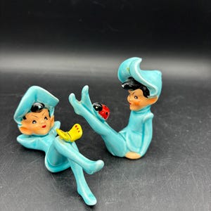 May include: Two ceramic figurines of elves in light blue outfits with black hair and rosy cheeks. One elf has a yellow butterfly on its leg, and the other has a red ladybug. The figurines are in a relaxed pose.