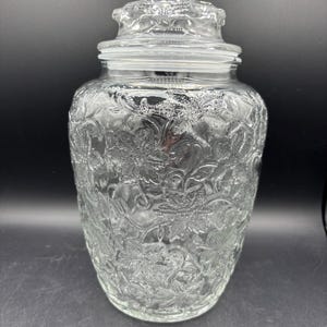 May include: Clear glass jar with a decorative floral pattern. The jar has a matching lid with a similar embossed design. The jar is cylindrical with a slightly wider base and a narrower neck. The glass is transparent, allowing the contents to be visible.