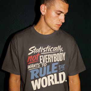 以下が含まれることがあります： 「Statistically, not EVERYBODY WANTS TO RULE THE WORLD.」というテキストがプリントされたチャコールグレーの半袖Tシャツ。「not」は赤、「RULE THE」は青、その他の文字は白で、太い筆致のフォントでデザインされています。