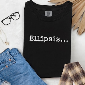 Puede incluir: Camiseta negra con la palabra "Ellipsis..." impresa en blanco. La camiseta está doblada y se muestra con gafas, un collar y unos vaqueros azules.