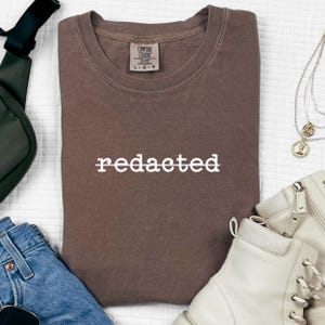 Könnte beinhalten: Ein braunes Comfort Colors Crew-Neck-Sweatshirt mit dem Wort "redacted" in weißen Blockbuchstaben. Das Sweatshirt liegt flach auf einer weißen Oberfläche, mit einer grünen Tasche, blauen Jeans, weißen Stiefeln und Schmuck in der Nähe.