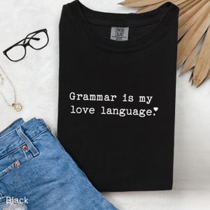 May include: Black t-shirt with the text "Grammar is my love language." in white. The shirt is folded and placed on a light-colored surface, with a pair of glasses and a necklace nearby. The shirt is made of a soft material.