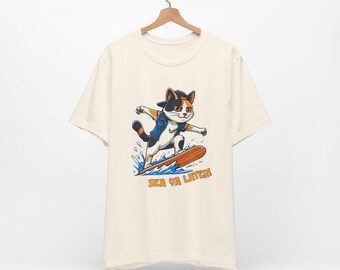 Puede incluir: Camiseta color crema con un gato de dibujos animados surfeando en una tabla de surf. El gato lleva una chaqueta vaquera y una gorra de béisbol. El texto "SEA YA LATER!" está impreso debajo.