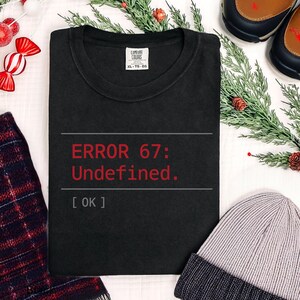 May include: Black t-shirt with the text "ERROR 67: Undefined." in red. The shirt has a crew neck and a tag that reads "COMFORT COLORS XL-TG-EG". A gray beanie, plaid scarf, and shoes are in the background.