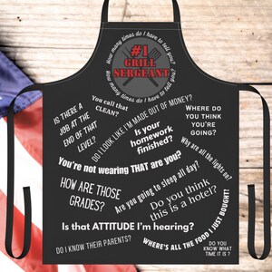May include: Black apron with the text "#1 GRILL SERGEANT" and various humorous phrases in white. The apron has a black neck strap and waist ties. The background includes a portion of an American flag.