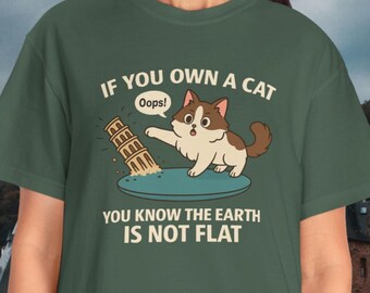 May include: Green t-shirt with the text "IF YOU OWN A CAT, YOU KNOW THE EARTH IS NOT FLAT." A cartoon cat is depicted pushing over a building. The cat is brown and white, and the building is beige.