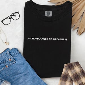 May include: Black t-shirt with the text "MICROMANAGED TO GREATNESS" in white. The shirt is folded and surrounded by accessories, including eyeglasses, a necklace, blue jeans, and a plaid scarf. The shirt has a Comfort Colors tag.