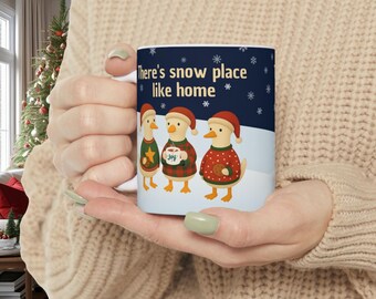 Op de afbeelding: Witte keramische mok met drie cartoon eenden in kersttruien en kerstmutsen. De mok heeft de tekst "There's snow place like home" en sneeuwvlokken. Een seizoensgebonden cadeau.