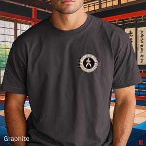 Pode incluir: T-shirt grafite com um emblema de Judo no peito. O emblema apresenta um praticante de Judo e as palavras "Judo Leverage Over Force" num design circular. A t-shirt é de manga curta.