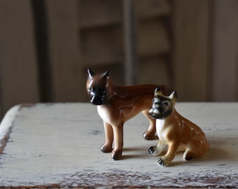 May include: Two ceramic Boxer dog figurines. One is standing, the other is sitting. Both have brown and black markings and are posed on a distressed white surface. The background is blurred, suggesting a vintage or antique setting.