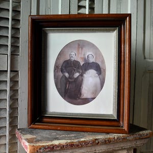 May include: A vintage oval portrait of a couple in a dark wood frame. The man is wearing a dark uniform and a hat. The woman is wearing a white dress and a white hat. The portrait is in a light brown oval mat.