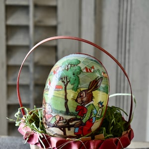 May include: An Easter egg with a painted scene of rabbits, trees, and houses, set inside a woven basket with a red handle. The egg is decorated with a vintage-style illustration and rests on green decorative grass.