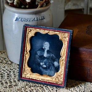 May include: Antique framed portrait featuring a seated woman in dark clothing. The portrait is in a decorative gold frame with a red border. The word "MUSTARD" is visible on a ceramic jar in the background.