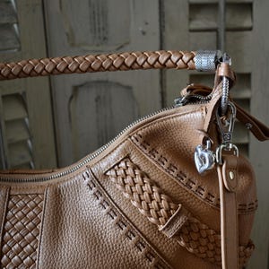 Pu&ograve; includere: Borsa a mano in pelle marrone chiaro con manico intrecciato e cuciture decorative. La borsa presenta una chiusura argentata e un ciondolo a forma di cuore. La pelle ha un aspetto strutturato.