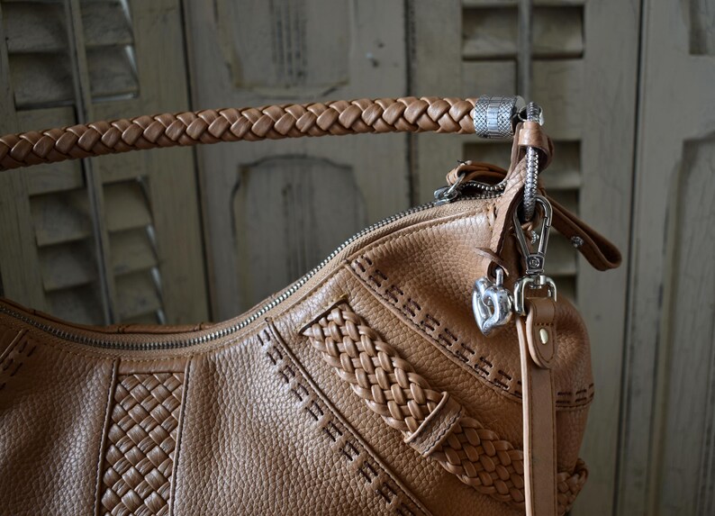 Pu&ograve; includere: Borsa a mano in pelle marrone chiaro con manico intrecciato e cuciture decorative. La borsa presenta una chiusura argentata e un ciondolo a forma di cuore. La pelle ha un aspetto strutturato.