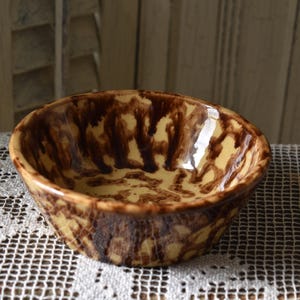 May include: A round ceramic bowl featuring a glossy finish with a cream-colored base and dark brown mottled, drip-like patterns covering both the interior and exterior surfaces. The bowl has a smooth, curved rim and a deep, rounded basin.