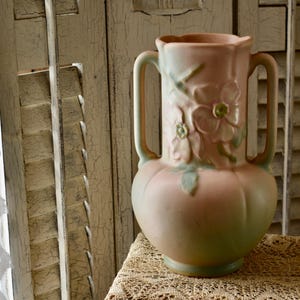 May include: A vintage, two-handled vase in shades of pink and green. The ceramic vase features embossed floral designs and a bulbous body. The handles are curved, and the vase sits on a lace-covered surface.