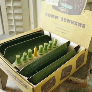 May include: Vintage corn server set in a cardboard box. The set includes green serving trays, corn holders, and a yellow corn cob holder. The box has text that reads "Corn Servers" and illustrations of the product.