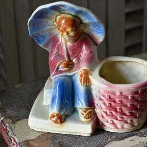 May include: A glazed ceramic figurine planter featuring a seated figure holding a blue and white umbrella over their head. The figure wears a red top and blue trousers, positioned next to a cylindrical basket-weave container with a pink finish.