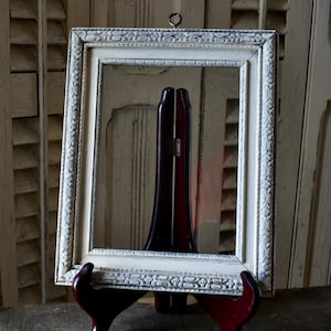 May include: A rectangular, off-white picture frame with ornate detailing, supported by a dark red easel. The frame has a decorative border and a small metal ring at the top for hanging. The easel is a dark red color.