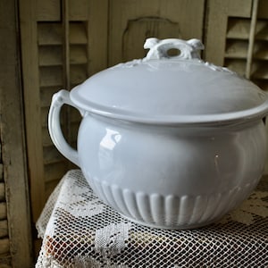 Peut inclure: Pot de chambre en céramique blanche avec un couvercle et une poignée décorative. Le pot a un design cannelé et est posé sur une nappe en dentelle blanche.