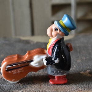 May include: A vintage wind-up toy of a man with a large nose wearing a blue top hat and a black suit holding a brown cello. The toy is made in Hong Kong and has the Walt Disney Productions copyright on the cello.