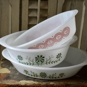 May include: Three white Pyrex casserole dishes stacked together. The top dish features a red and white pattern, while the middle dish has a green floral design. The oval dishes have handles. The dishes are stacked on top of each other.