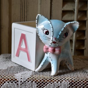 Puede incluir: Un macetero de cerámica azul y blanco con forma de gato con una pajarita rosa. El macetero tiene forma de bloque con la letra "A" en rosa en la parte delantera.