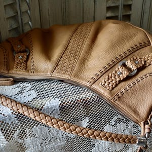 Pu&ograve; includere: Borsa a tracolla in pelle color cuoio con cinturino intrecciato e cuciture decorative. La borsa presenta un pannello intrecciato e finiture color argento. La cerniera &egrave; aperta, rivelando l'interno. La borsa &egrave; appoggiata su una superficie di pizzo bianco.