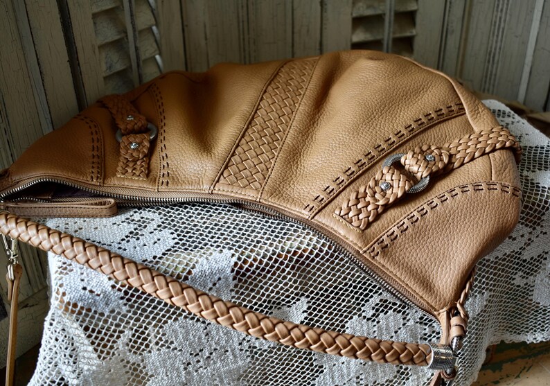 Pu&ograve; includere: Borsa a tracolla in pelle color cuoio con cinturino intrecciato e cuciture decorative. La borsa presenta un pannello intrecciato e finiture color argento. La cerniera &egrave; aperta, rivelando l'interno. La borsa &egrave; appoggiata su una superficie di pizzo bianco.