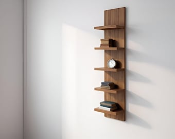 May include: A vertical wooden wall shelf with five horizontal shelves. The shelves hold books and a small clock. The wood has a warm, natural tone, and the shelf is mounted on a white wall.