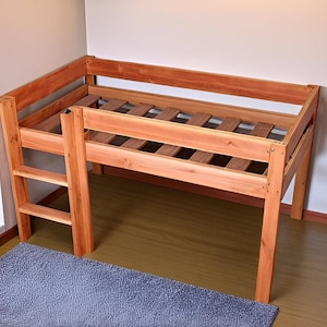 May include: A wooden loft bed with a natural finish. The bed features a ladder for access and a slatted base. The frame is made of solid wood and has a simple, sturdy design. The bed is positioned in a corner of a room, with a gray rug below.