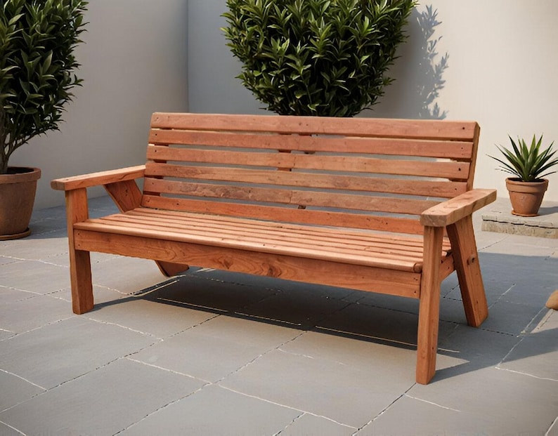K&ouml;nnte beinhalten: Eine Holzbank mit nat&uuml;rlichem Finish. Die Bank hat eine Lattenlehne und -sitzfl&auml;che mit Armlehnen. Die Bank ist etwa 152 cm lang und aus Holz gefertigt. Die Bank befindet sich im Freien auf einer Terrasse.