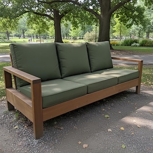 May include: A wooden outdoor sofa with three olive green cushions and backrests. The sofa has a natural wood frame and is placed on a gravel surface in a park setting. The sofa has a modern design.