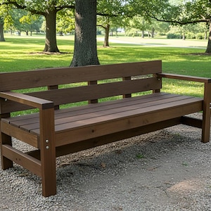 May include: A brown wooden park bench with a slatted seat and backrest. The bench has armrests and is constructed with square posts and supports. It is set on a gravel surface, with a green lawn and trees in the background.