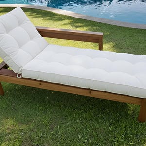 May include: A wooden chaise lounge with a white tufted cushion. The frame is made of brown wood, and the cushion is a light cream color. The lounge is on a grassy lawn near a pool.