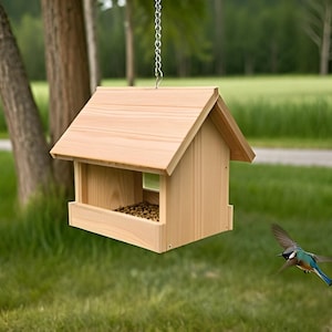 May include: A wooden bird feeder with a pitched roof, hanging from a chain. The feeder is filled with birdseed and a small bird is flying towards it. The background includes trees and green grass.