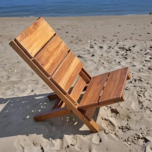 Peut inclure: Chaise de plage pliante en bois avec dossier et assise à lattes. La chaise est en bois brun clair et est posée sur une plage de sable avec l'océan en arrière-plan. La chaise est inclinée, montrant son design.