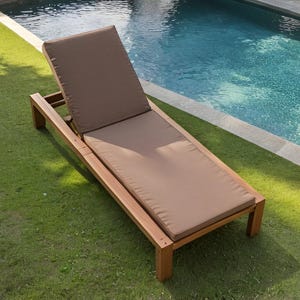 May include: A wooden chaise lounge with a brown cushion and backrest sits on green grass next to a pool. The lounge chair is made of light-colored wood and has a rectangular shape.