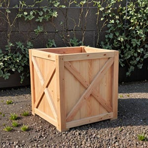 Könnte beinhalten: Ein großer, quadratischer Pflanzkasten aus hellem Holz. Die Box hat ein X-förmiges Design auf jeder Seite für zusätzliche Stabilität und optisches Interesse. Der Pflanzkasten ist leer und bereit für Pflanzen. Die Box steht auf einem Bett aus kleinen Steinen.