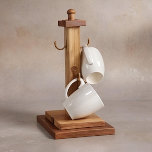May include: A wooden mug tree with three curved hooks, holding two white ceramic mugs. The tree has a square base and a cylindrical top. The wood has a natural finish.