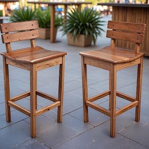 May include: Two wooden bar stools with a natural finish. Each stool has a backrest with two horizontal slats and a contoured seat. The legs are square and connected by a footrest. The stools are made of light brown wood.