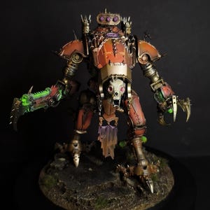 May include: A detailed miniature robot figure with a skull-like face and clawed hands. The figure is primarily orange and gold, with green and purple accents. It stands on a textured, dark base.