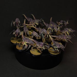 Puede incluir: Colección de criaturas en miniatura con forma de insectos, con exoesqueletos morados y verdes, posadas sobre una base negra con una superficie texturizada y beige. Las figuras están dispuestas en un grupo, con garras afiladas y detalles.