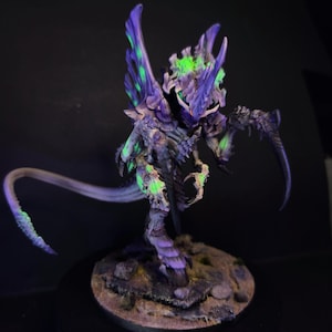 Könnte beinhalten: Detaillierte Miniaturfigur einer Kreatur mit lila und grüner Farbgebung. Die Figur hat einen langen Schwanz, scharfe Krallen und einen strukturierten Körper. Sie steht auf einer kleinen, strukturierten Basis mit dunklem Hintergrund.