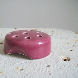 May include: A small, pink ceramic flower frog with seven holes for holding stems. The frog has a slightly irregular shape and a glossy finish.