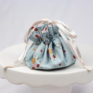 May include: A small, light blue drawstring bag with a floral pattern of red and yellow flowers and green leaves. The bag is tied with a cream-colored ribbon and sits on a white, scalloped-edge cake stand.